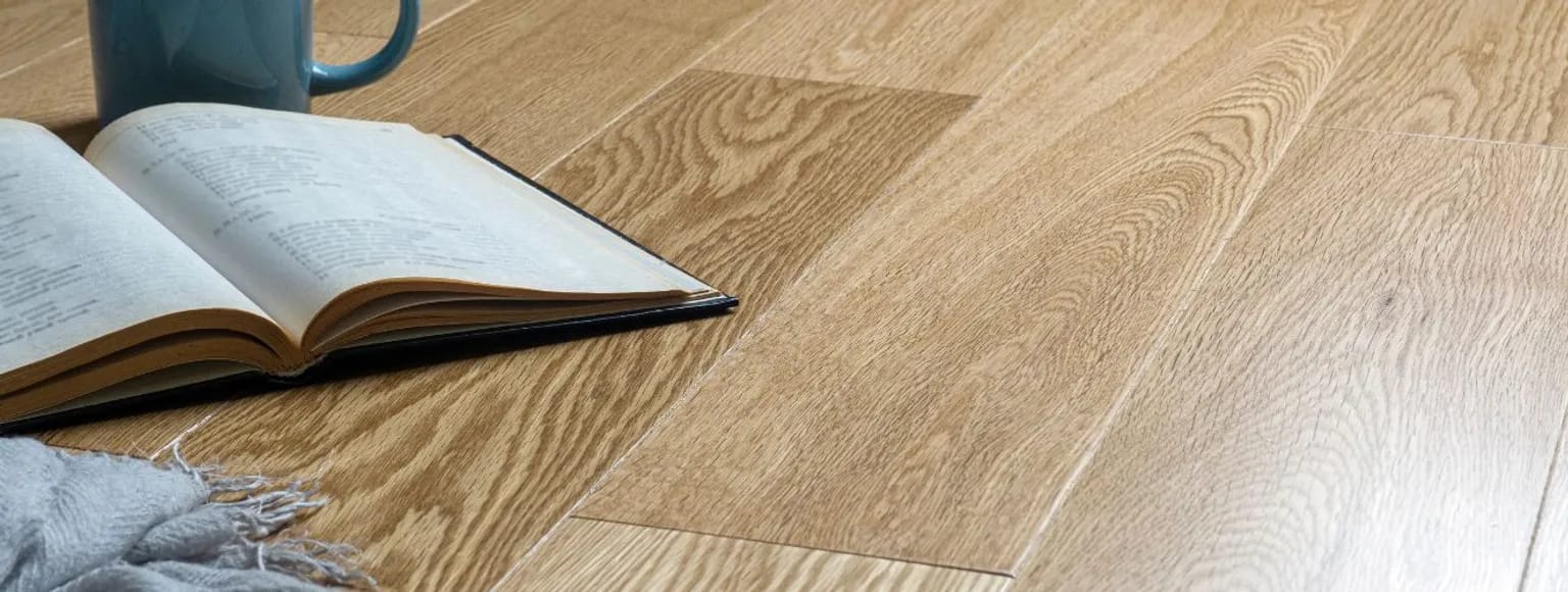 Aufgeschlagenes Buch, blauer Becher und graues Tuch auf hellbraunem XXL-Holzdielenboden.