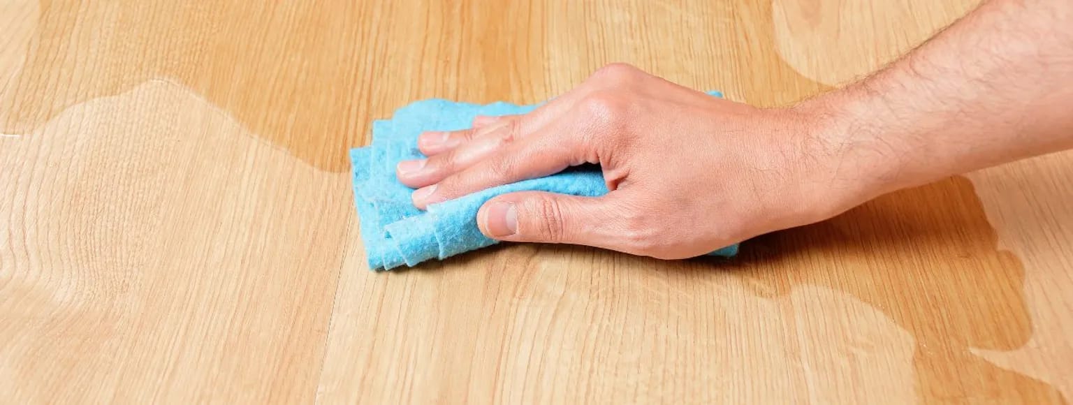 Eine Hand wischt verschüttete Flüssigkeit von einem hellen Holzboden und veranschaulicht so die Schritte der Parkettpflege.