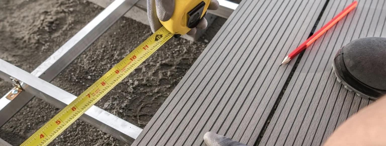 Handschuhe benutzen ein Maßband auf grauen WPC-Terrassendielen, ein roter Bleistift liegt in der Nähe.