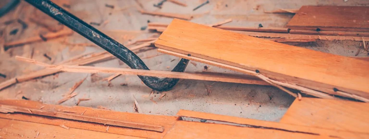 Mit einem metallenen Brecheisen wird der alte Bodenbelag entfernt, wobei sich Holzsplitter auf dem hellen Boden verteilen.