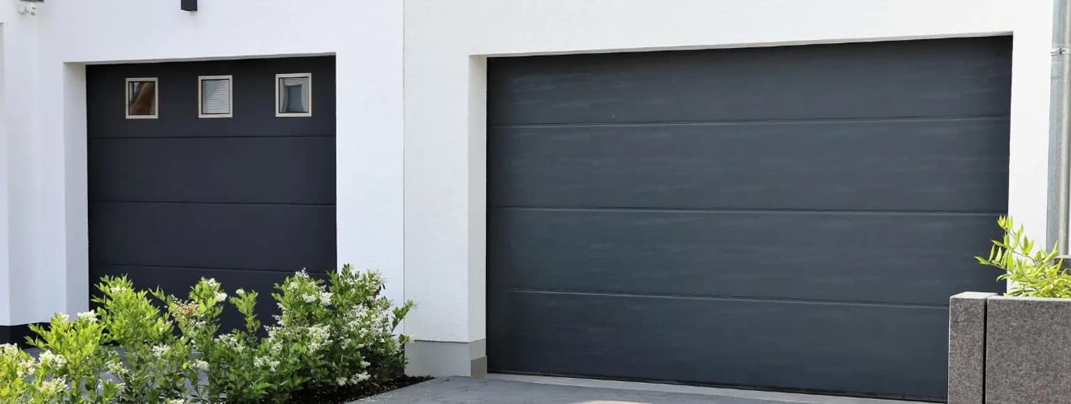 Schwarze Garagentore mit horizontalen Linien an einem weißen Haus, davor grüne Sträucher.