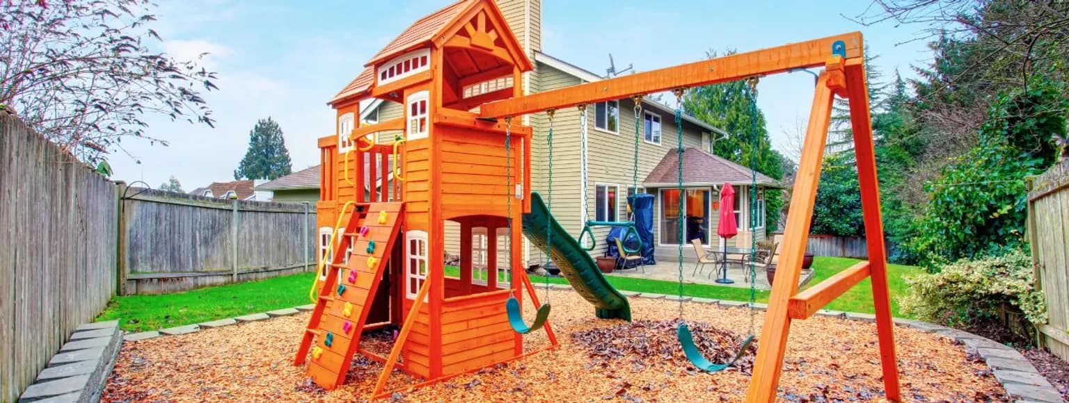 Spielplatz aus Holz mit Schaukeln und Rutsche in einem Garten, umgeben von Gras.