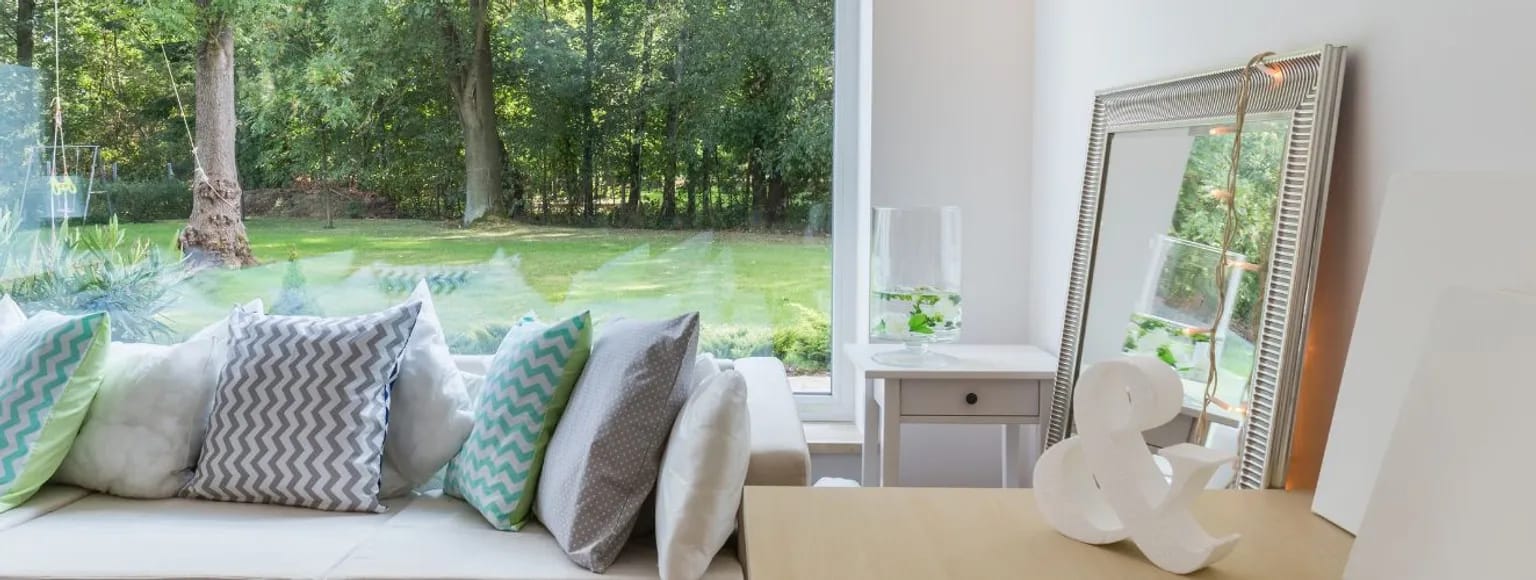 Wohnzimmer mit gemusterten Kissen, weißen großen PVC-Fenstern und Blick in einen grünen Garten.