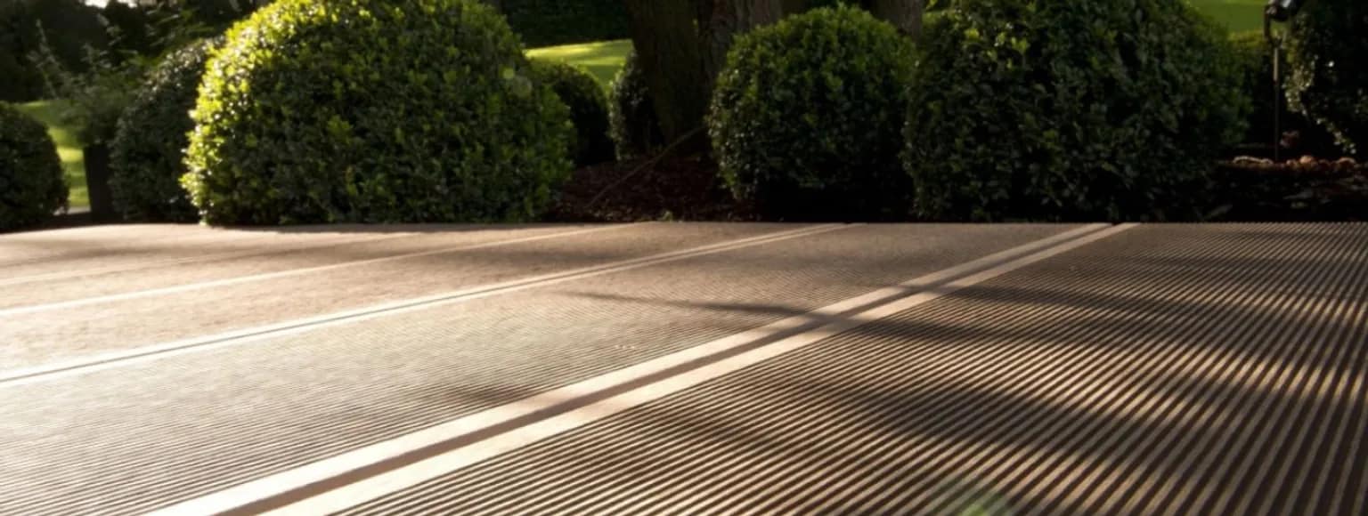 WPC-Terrassenreinigung im Sonnenlicht, mit parallelen Rillen und beschnittenen grünen Büschen dahinter.
