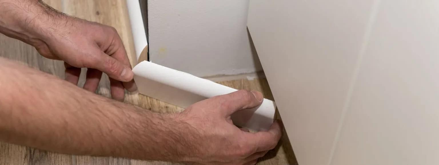 Zwei Hände, die eine weiße Sockelleiste entlang der Kante eines hellen Holzfußbodens im Innenbereich anbringen.