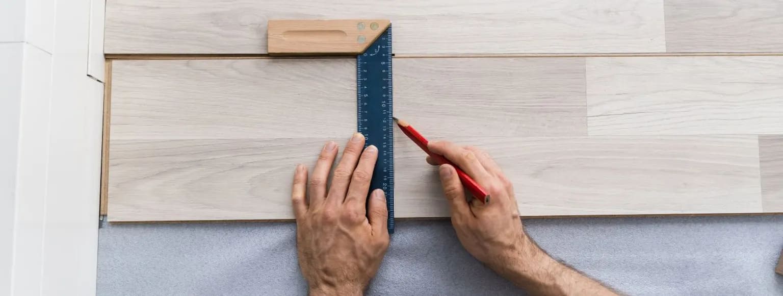 Zwei Hände messen den hellbraunen Laminatboden mit einem Lineal während der Verlegung aus.