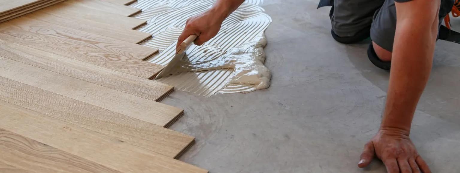 Zwei Hände verteilen Klebstoff auf einem grauen Betonboden und verlegen helle Holzdielen.
